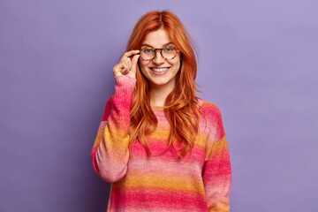 Young pretty redhead woman corrects glasses and wears sweater poses against purple background. Adorable glad foxy lady being in great mood stands indoor. Charming teenager has pleased expression