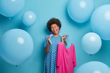 Positive overjoyed young African American woman chills indoor, holds high heel shoe as microphone and chooses best outfit to wear. Glad stylish lady carries fashionable pink dress on hangers