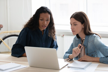 African American businesswoman mentor training intern, using laptop, sitting at table in modern...