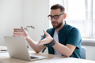 Confident businessman wearing glasses making video call, using laptop, looking at screen, sitting at work desk in modern office, successful young man worker consulting client, chatting online