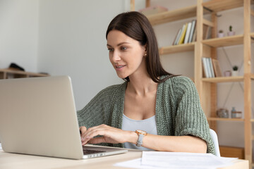 Smiling young businesswoman using laptop, sitting at desk in modern office, confident focused employee intern looking at computer screen, working on online project, writing email, typing