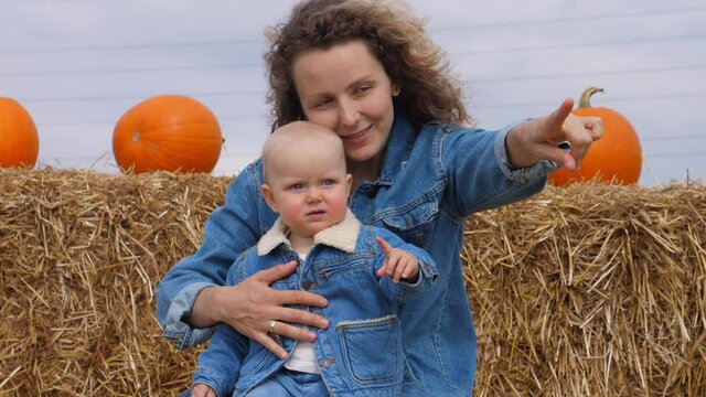 Portrait of Caucasian mom and baby in matching denim looks sitting on a hay bale pointing at something in the distance. Special bond between young single mother and a child