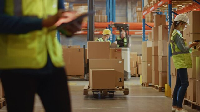 Male Worker Wearing Hard Hat Checks Products Stock and Inventory with Digital Tablet Standing in Retail Warehouse full of Shelves with Goods. Arc Shot Moves to People Operating Forklifts and Trucks