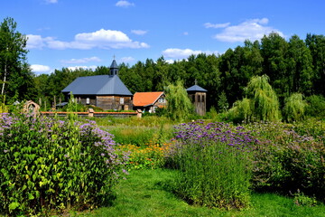 View of an old wooden Catholic church with an angled roof located next to a well maintained garden and a dense forest or moor seen on a cloudy yet warm summer day on a Polish countryside 