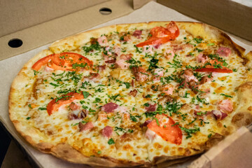Round pizza with ham, cheese, tomatoes and herbs and bacon in a cardboard box. Fast food delivery