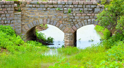 Fototapeta na wymiar Old stone single arch bridge, Latvia. Famous ancient stone arch single track road bridge in the forest. View of small river and trees reflection in the water.