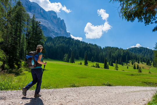 Young woman with a hat, trekking poles, a backpack and a spare face mask attached to it, hiking on a trail through green meadows in the Italian Alps. Dolomite peaks are visible in the background