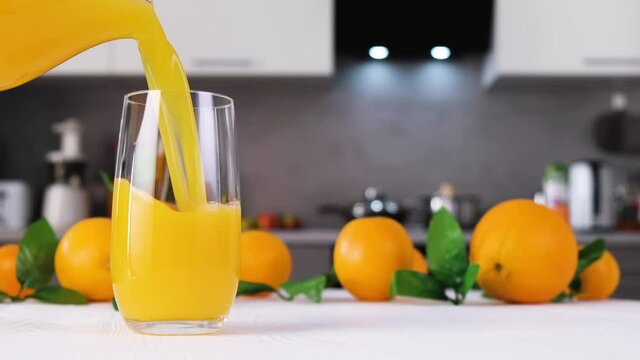 Orange juice pouring into glass on white table in modern kitchen. Citrus juice splash slow motion footage