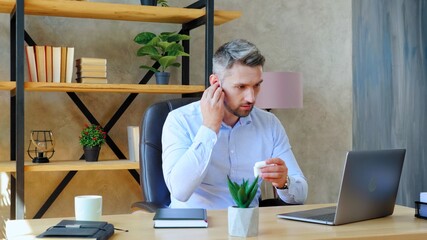 Gray-haired bearded business man in home office puts on wireless earphones using laptop computer. Guy connects to remote online video call chat conference with teacher, distance education