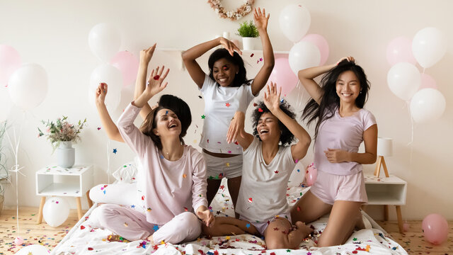 Energetic young diverse multiethnic girls best friends in sleepwear playing with colorful confetti on bed, entertaining at sleepover party, celebrating birthday or having fun together in bedroom.