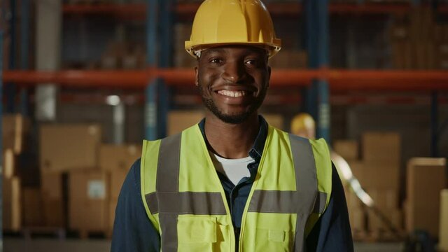 Handsome and Happy Professional Worker Wearing Safety Vest and Hard Hat Charmingly Smiling on Camera. In the Background Big Warehouse with Shelves full of Delivery Goods. Slow Motion Medium Portrait