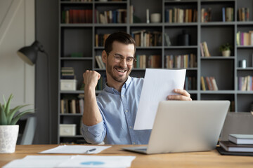 Excited young Caucasian man work on computer feel euphoric reading paper letter correspondence....
