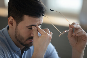 Close up of tired young man take off glasses massage eyes suffer from dizziness or blurry vision....