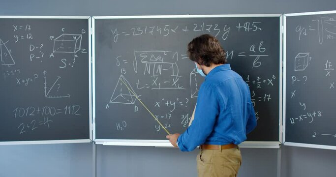 Caucasian handsome male teacher in medical mask standing at blackboard with formulas, graphics and laws and explaining with pointer. Lecturer of mathematics at school or college. Coronavirus concept.