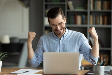 Overjoyed millennial male worker in glasses look at laptop screen triumph get pleasant promotion...
