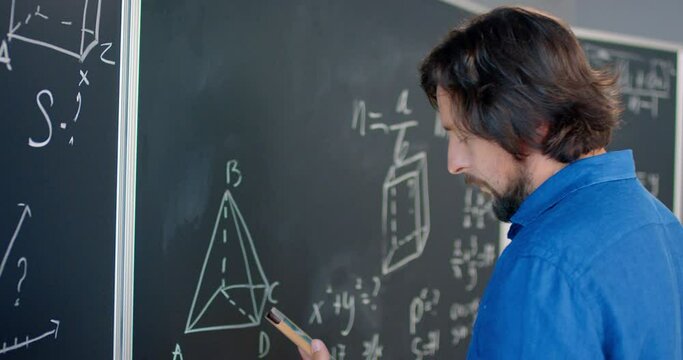 Close up of Caucasian male teacher in glasses writing formulas and math laws on blackboard and looking at tablet device. Educative mathematics lection. Man lecturer using gadget computer as cribe.