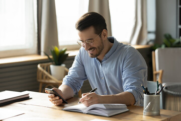 Smiling young Caucasian man sit at desk at home office workplace look at cellphone screen watch webinar making notes. Happy millennial male use smartphone summarize plan writing in journal.