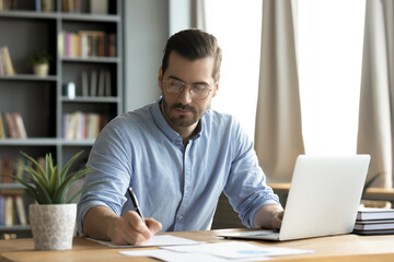Concentrated young caucasian man in glasses sit at desk work on computer make notes. Focused...