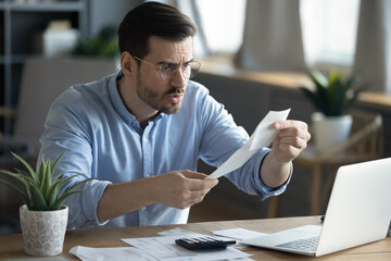 Angry millennial Caucasian guy sit at desk managing financial paperwork frustrated by bill amount....