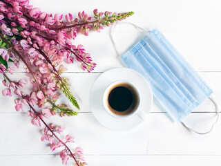 Cup of coffee, removed medical mask and pink flowers. Breakfast in the cafe. Modern reality and life concept. Blue surgical protective mask and white cup on white wooden background. Flat lay, top view