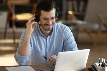 Smiling millennial Caucasian man in headset look at laptop screen work online from home office....