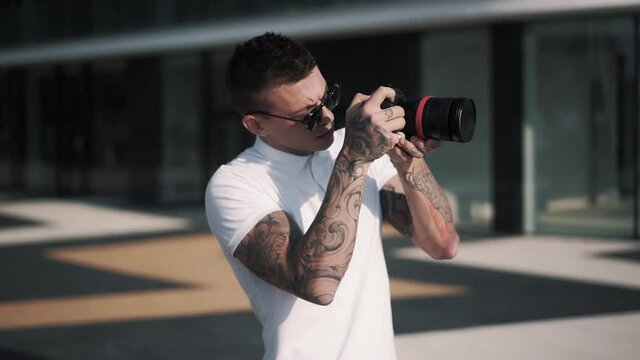 Tattooed male photographer in sunglasses holds camera in hands and takes photo