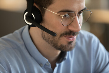 Crop close up of young Caucasian man in headphones glasses consult client or customer online. Millennial male employee in headset talk speak using wireless internet connection. Technology concept.