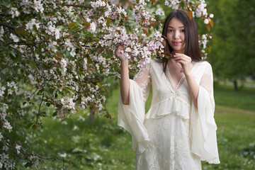 A beautiful Asian woman in a white dress, standing near a tree with white flowers and holding branches with her hands, looks directly into the camera