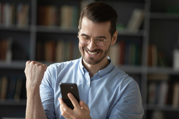 Overjoyed young Caucasian man in glasses look at smartphone screen feel euphoric win lottery...