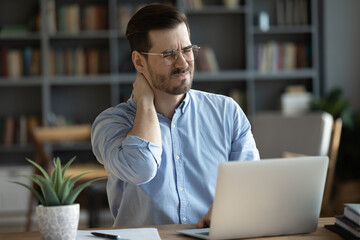 Exhausted young Caucasian male worker sit at desk massage neck suffer from strain spasm muscles....