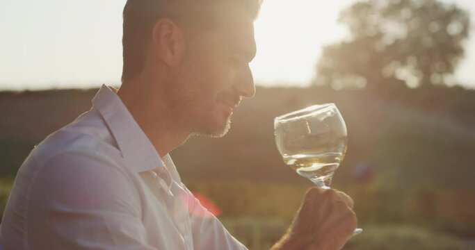 Authentic shot of happy handsome elegantly dressed man is tasting a flavor and drinking fresh white wine poured in transparent glass on scenic vineyards background at sunset.