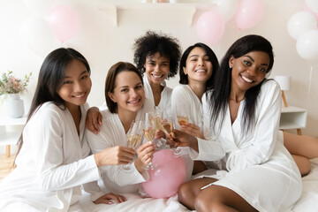 Portrait of smiling young mixed race pretty female best friends in white silk gowns, enjoying bachelorette hen pre-wedding party with sparkling wine, toasting saying wishes at luxury spa celebration.