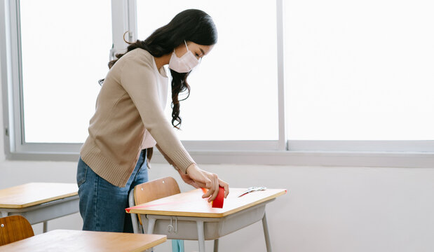 Asian Teacher In A Medical Mask Marking Places In The Classroom For Returning Of Students After Covid19 Quarantine And Lockdown. Social Distancing. Focus Hand.