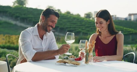 Authentic shot of happy couple in love is enjoying romantic dinner together and cheering with white wine glasses to celebrate their anniversary and timeless love on scenic vineyards background.