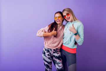 Two girls with surprise face  staying  over purple wall. Wearing fashionable hoodies and cool glasses.