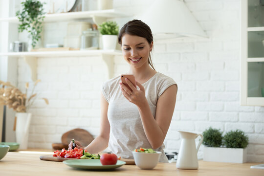 Smiling Young Woman Making Salad And Using Mobile To Count Calories And Plan Diet. Happy Housewife Consulting Recipe Instructions On Smartphone While Chopping Vegetables In Modern Kitchen