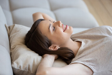 Obraz na płótnie Canvas Happy relaxed woman lying on couch with eyes closed and enjoying peace and quiet of her own soundproof house. Good-looking young lady lounging on sofa cushion and smiling feeling content and fulfilled