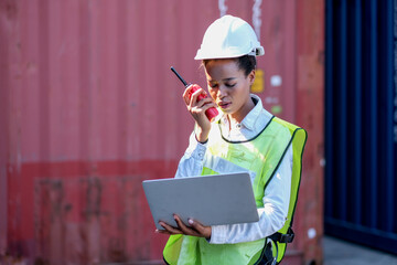 American African factory worker woman talk to red walkie-talkie and laptop for her working in container shipping area. Concept of good system and manager support for better industrial business.