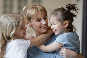 Close up of happy young mother in casual wear cuddling her two little daughters and smiling joyfully. Cute children embracing their mommy to express love and affection