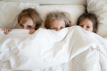 Happy mom and children lying on comfortable bed together and peeking out from under soft white comforter. Young mother and daughters hiding half-face under warm duvet looking up at camera. Top view