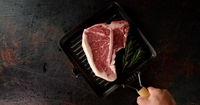 A frying pan with raw beef t-bone steak is put on table. 