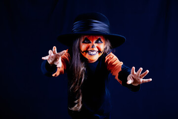 girl with pumpkin makeup and black hat scares and pretends to be a monster on a black background, Halloween celebration.