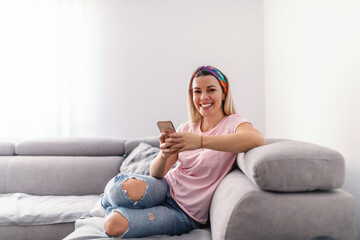 Young smiling gorgeous housewife sitting on couch in living room, taking a break and using smart...