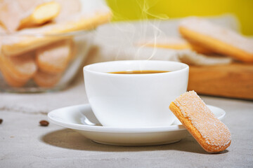 A white cup of hot aromatic coffee with steam and traditional italian savoiardi biscuits or ladyfingers cookie on a saucer and in a glass bowl. Selective focus
