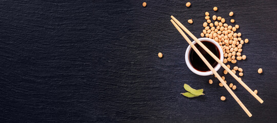 Soy sauce in a bowl with bamboo chopsticks and soya beans scattered on the black surface of the...