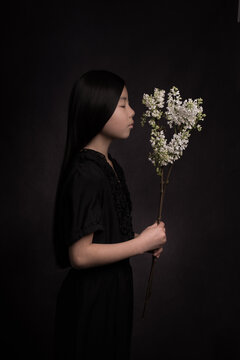 Teenage girl in black dress holding white lilac flowers while standing indoors
