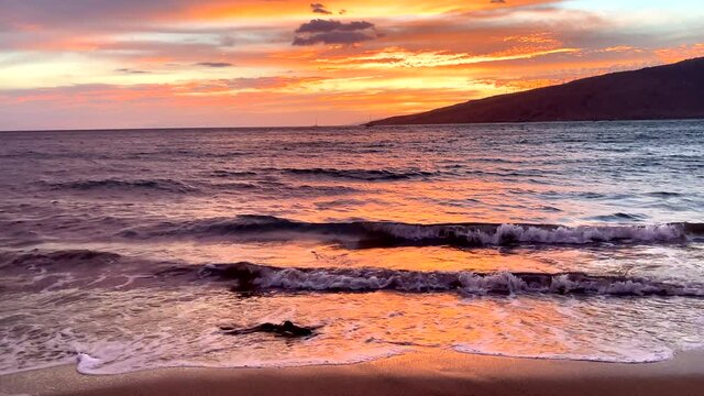Colorful Hawaii sunset with small rooms of surf playfully rolling small driftwood along shore with silhouette of part of island in distance
