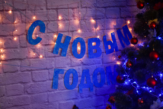 Happy new year in Russian on a brick background next to a Christmas tree lit with a garland