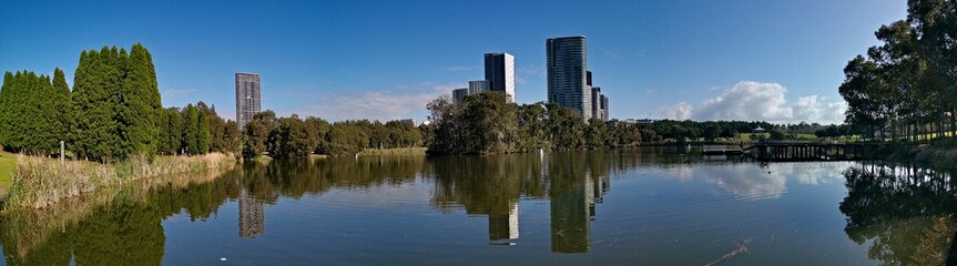 Fototapeta na wymiar Beautiful panoramic view of a lake with reflections of luxury high-rise building, blue sky, clouds, and trees on water, lake Pavillion, Sydney Olympic park, Sydney, New South Wales, Australia 