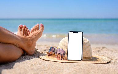 Smartphone with blank white display on the beach. Hat, sunglasses and legs of the girl on the...